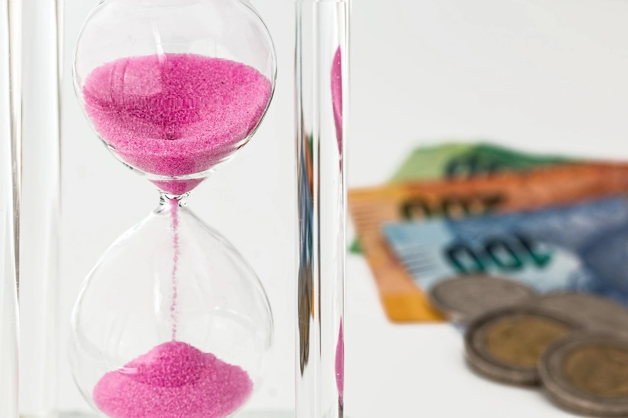 Reloj de arena, al fondo se ven monedas y papel moneda.